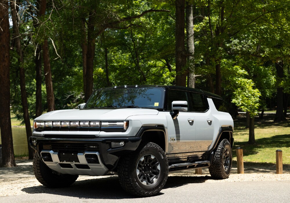 hummer-ev-for-sale.png Photo of white 2024 Hummer SUV EV parked in front of several trees. The Hummer logo is visible on the front grilled of the car