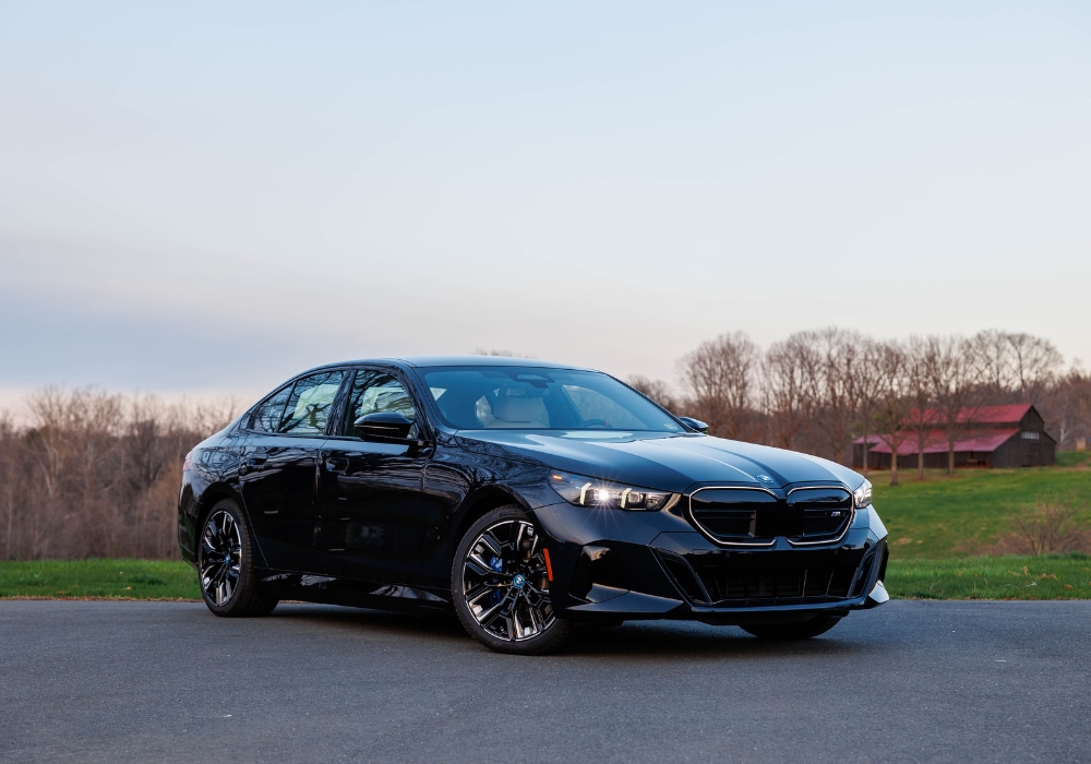 2024-bmw-i5.png Black 2024 BM i5 parked in front of a tree line with a barn out of focus in the background