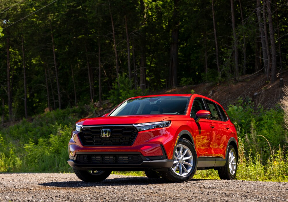 honda-cr-v-for-sale-near-me.png 2025 Honda CR-V parked on a gravel road with greenery in the background. The SUV is bright red and the Honda logo is visible on the front grille