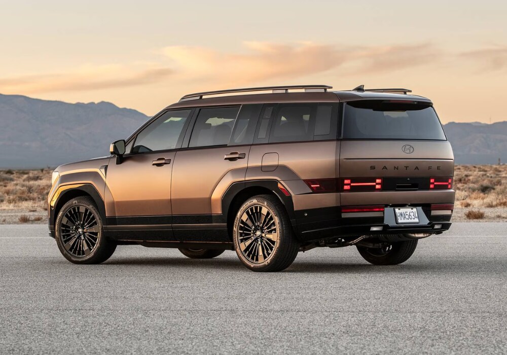 hyundai-santa-fe-specs.png A 2025 Hyundai Santa Fe parked in a lot in front of a mountain range at sunset. The Santa Fe logo visible on the back of the car and its taillights are on