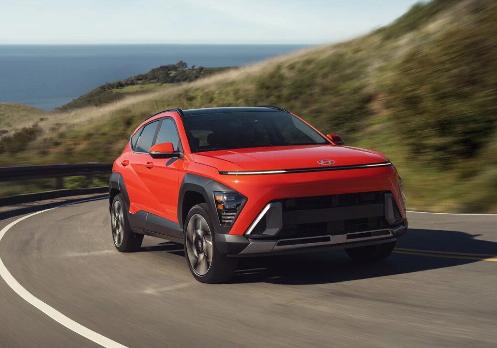 hyundai-kona-for-sale.png Orange 2024 Hyundai Kona driving down a paved road, facing the camera