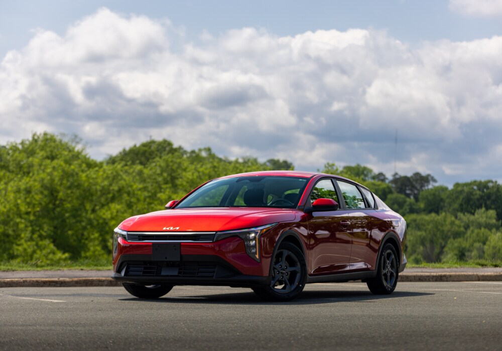 kia-k4-2025.png 2025 Kia K4 parked on a paved lot with trees in the background. The car is bright red and the front of the vehicle is facing the camera