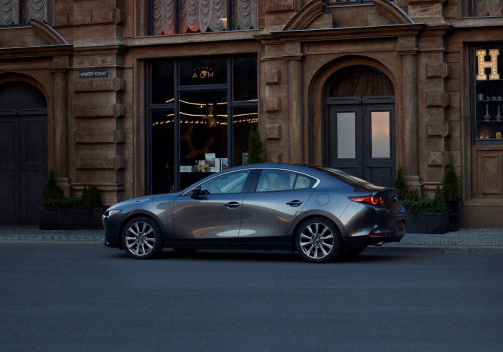 mazda3-sedan-2025.png Dark gray Mazda3 parked in front of a stone building