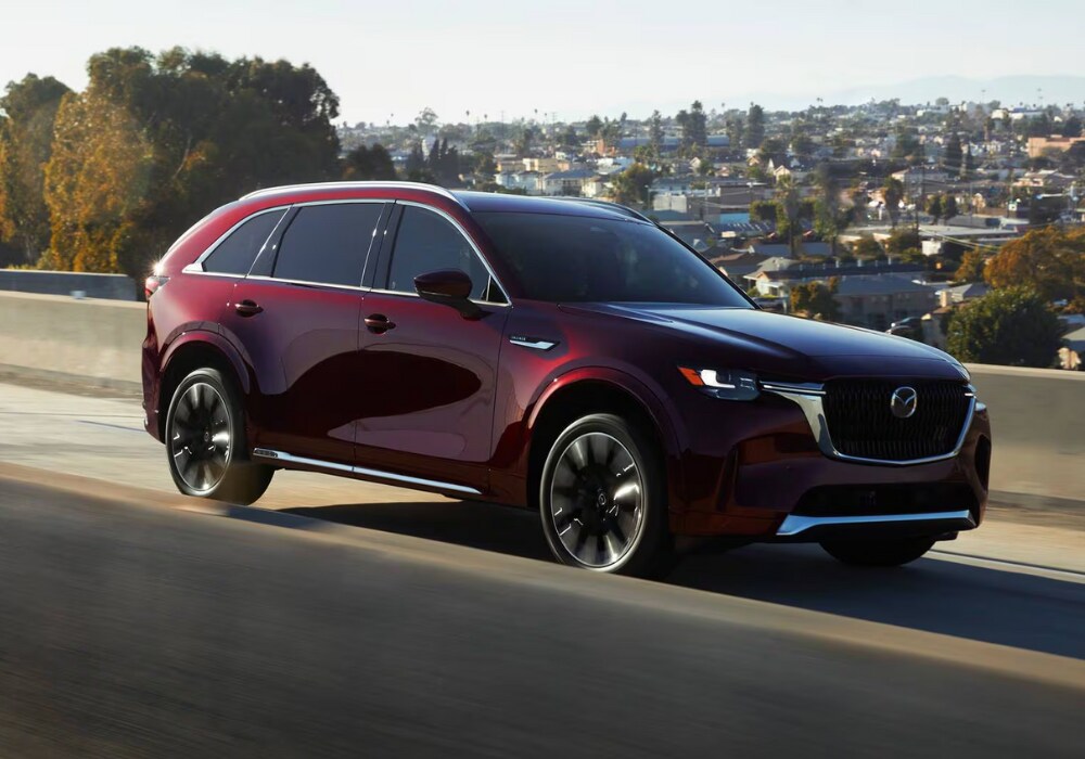 mazda-cx-90-for-sale.png Red 2025 Mazda CX-90 driving with a city in the background