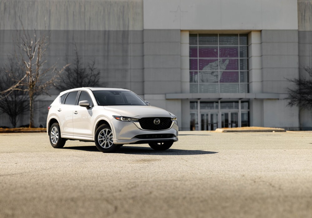 new-mazda-cars-for-sale-fayetteville.png White Mazda CX-5 SUV for sale parked in an empty lot. There is a large building in the background