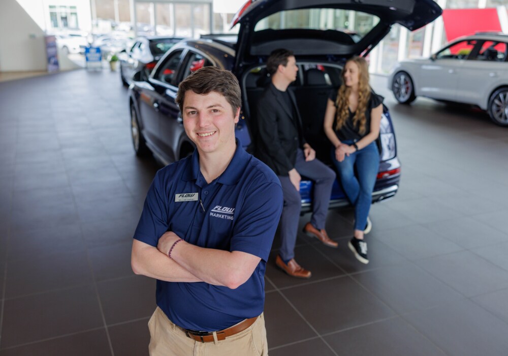 used-mazda-for-sale-greensboro.png A Flow sales person smiles and looks at the camera while two customers rest against the trunk of an SUV in the background
