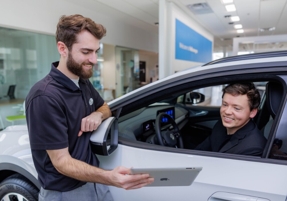 used-volkswagen-for-sale-greensboro.png A Flow employee assisting a customer who is sitting inside a car at Flow Volkswagen dealership. The employee is smiling and showing the customer an iPad