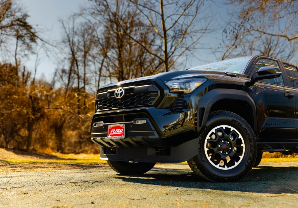 toyota-tacoma-for-sale.png The front half of a Toyota Tacoma truck parked on a paved lot with trees in the background. The truck is black and the front plate has the Flow Automotive logo on it