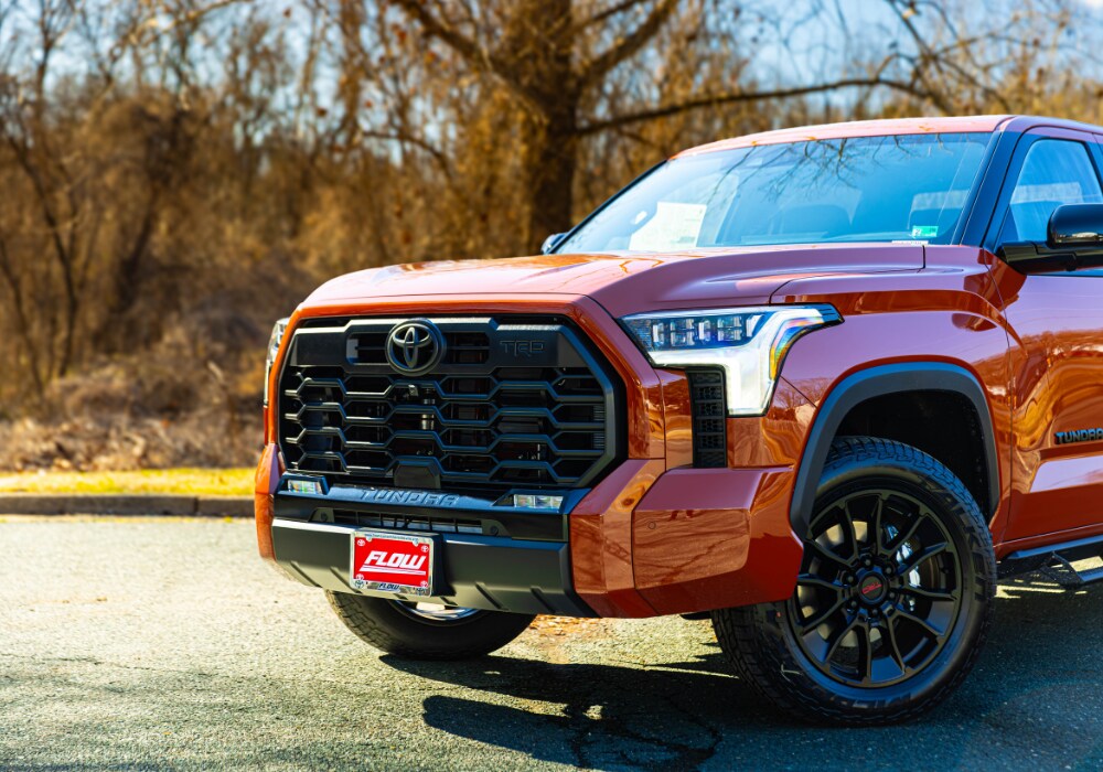 toyota-tundra-for-sale.png The front half of a Toyota Tundra truck parked in a Charlottesville parking lot. The truck is orange, its headlights are on, and the Flow Automotive logo is visible on the front plate of the truck