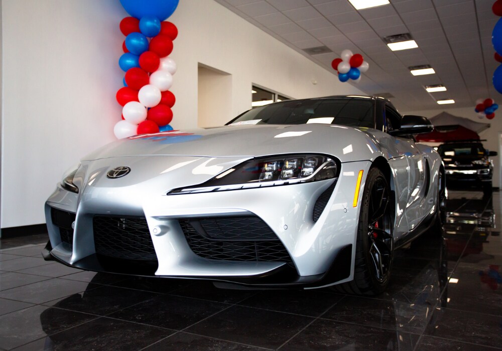 new-toyota-cars-for-sale-statesville.png A Toyota car parked inside the showroom of Flow Toyota of Statesville. There a red, white, and blue balloons in the background