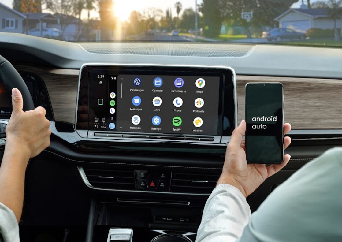 volkswagen-android-auto.png The front dash of a Volkswagen car with a smart display with various apps. A hand is holding a phone with Android Auto open on the screen