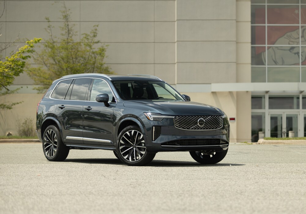 2025-volvo-xc90-for-sale.png Black 2025 Volvo XC90 SUV parked in a paved lot with a larger building in the background