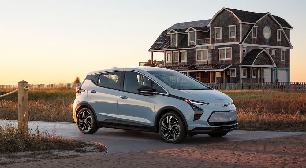 Roseville Chevy Dealers - 2022 Bolt EV - Driveway.jpg A silver 2022 Bolt EV parked near a large coastal home surrounded by grassy fields