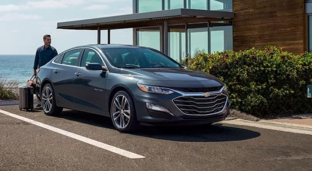 Roseville Chevy Dealers - 2022 Chevy Malibu - Grey (1).jpg A dark grey 2022 Chevy Malibu parked in front of a modern home overlooking the ocean