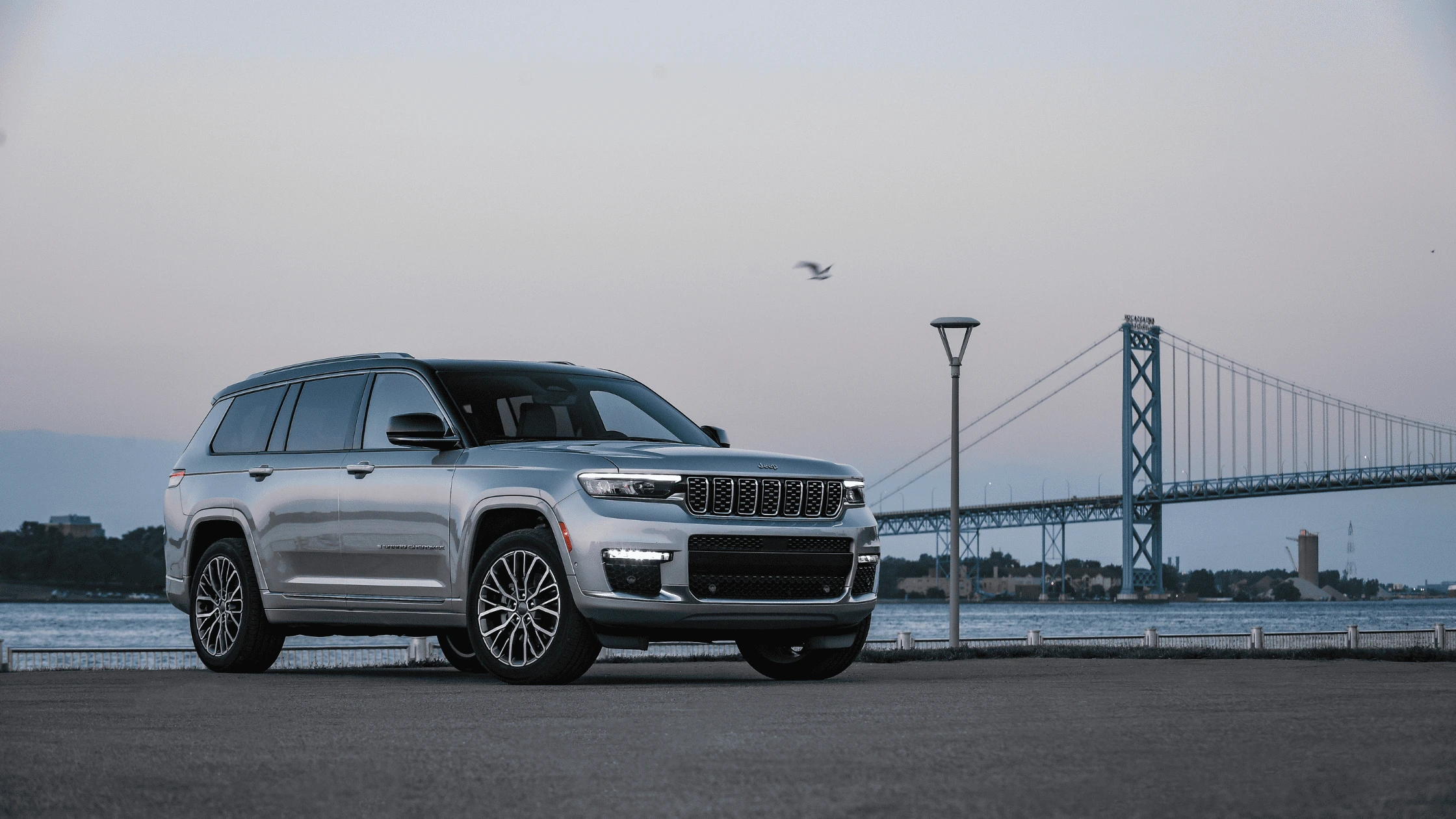 2025 Jeep® Grand Cherokee L in Fort Myers, FL Silver 2025 Jeep Grand Cherokee L by a bridge with water and skyline in background