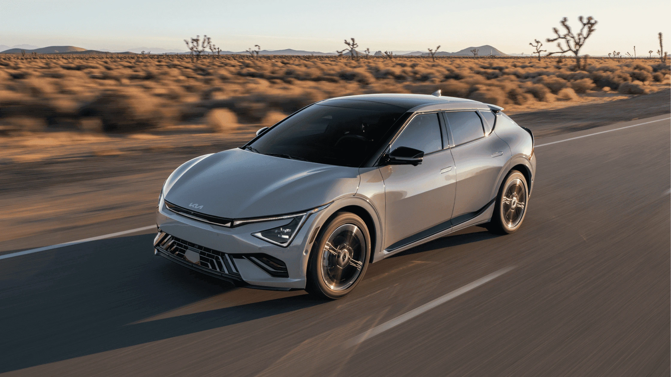 The 2025 Kia EV6 in Fort Myers, FL A silver 2025 Kia EV6 electric vehicle driving through a desert highway, showcasing its aerodynamic design, futuristic headlights, and sporty alloy wheels under the warm glow of the setting sun.