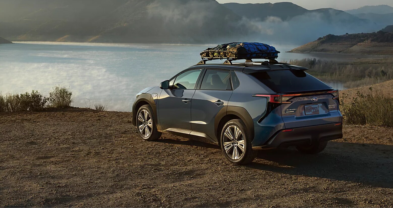 2023-Subaru-Solterra 2.png A new two-tone Harbor Mist Grey Subaru Solterra with camping supplies strapped on top parked on the shore of a cloudy lake