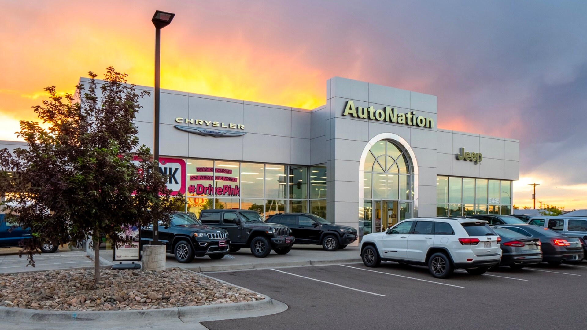 Exterior view of Autonation Chrysler Jeep Broadway during a sunrise or sunset. Many vehicles parked near the grey building. Trees and greenery are visible around the parking lot and surrounding area.