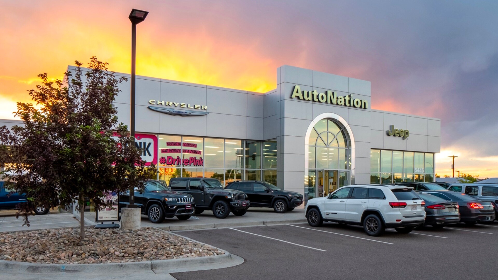 Autonation Chrysler Jeep Broadway is your Littleton, CO home for all your Chrysler, Jeep financing, purchasing, and service needs Exterior view of Autonation Chrysler Jeep Broadway during a sunrise or sunset. Many vehicles parked near the grey building. Trees and greenery are visible around the parking lot and surrounding area.