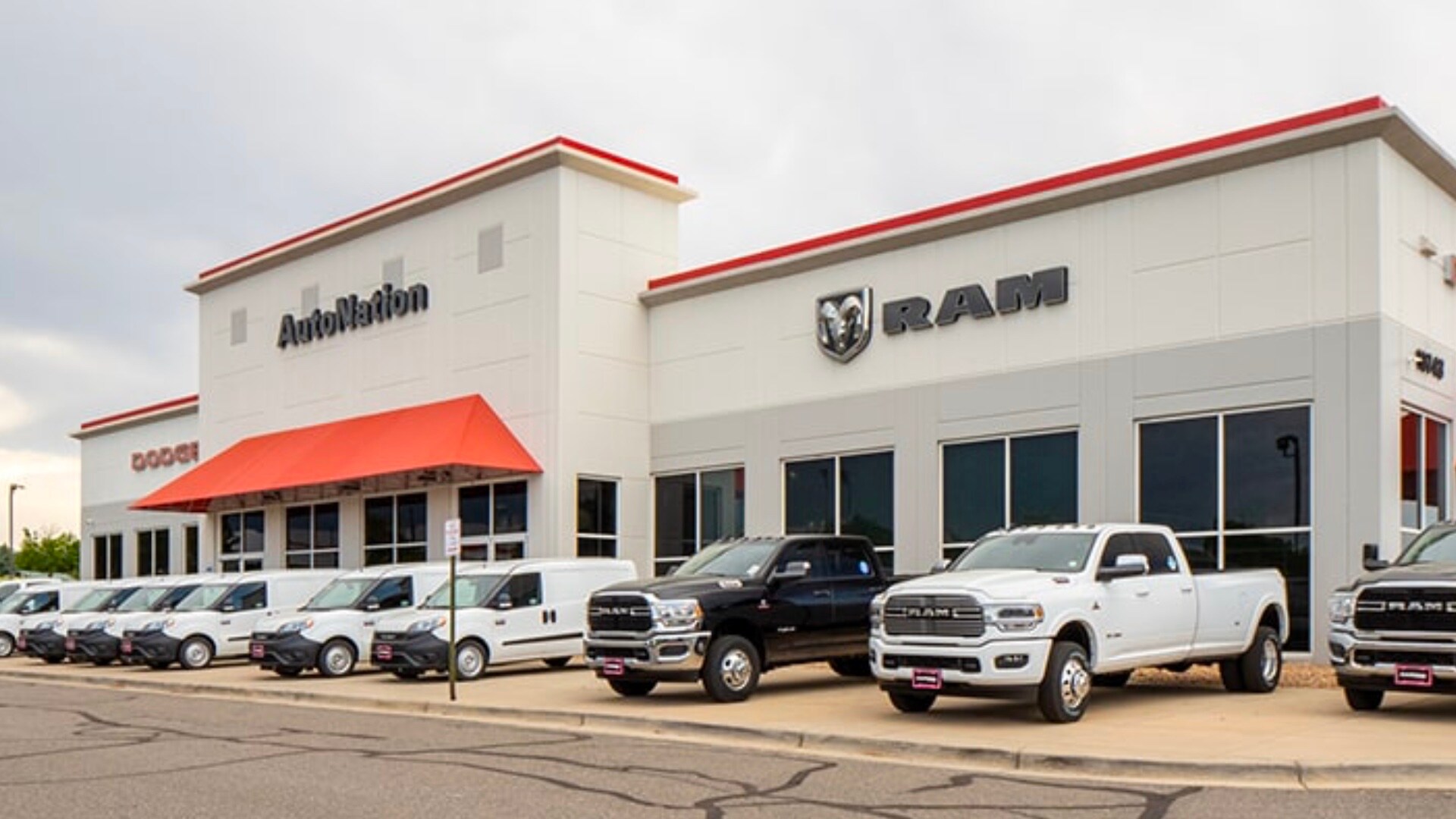 Exterior view of AutoNation Dodge Ram Arapahoe. There is a blue sky and many vehicles parked in front of the grey building. Some trees can be seen in the background.
