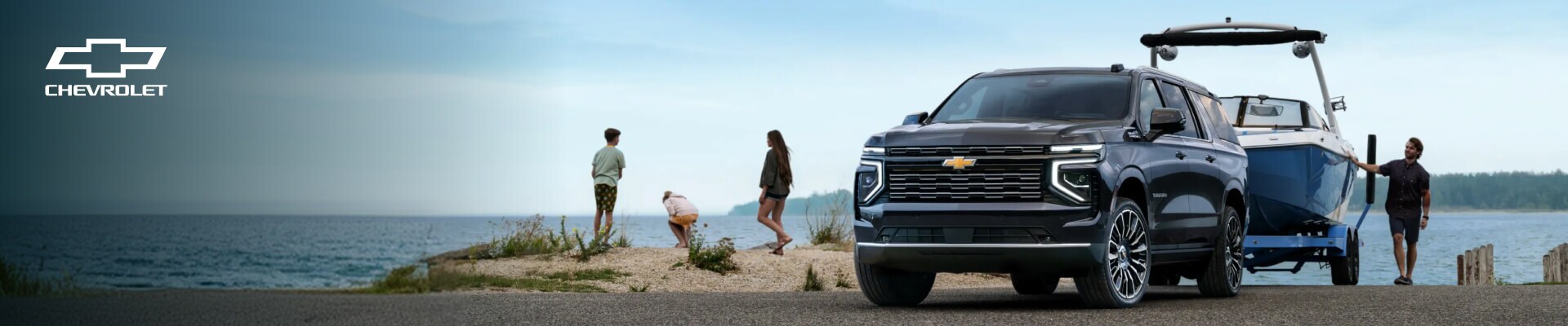 2026 Chevrolet Suburban High Country in Dark Ash Metallic towing a boat beside a lake with a family.