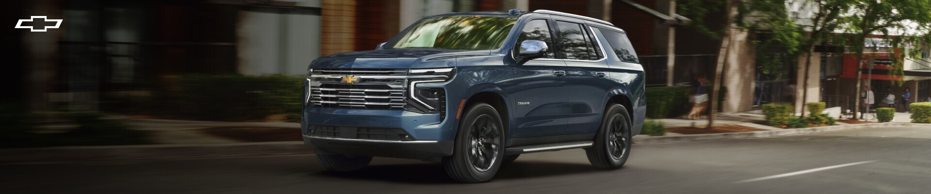 Chevrolet Tahoe accelerates past a modern building on a sunny city street.