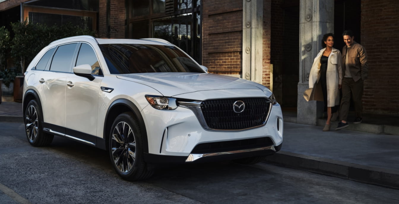 White Mazda CX-90 parked on a city street with two people walking nearby.