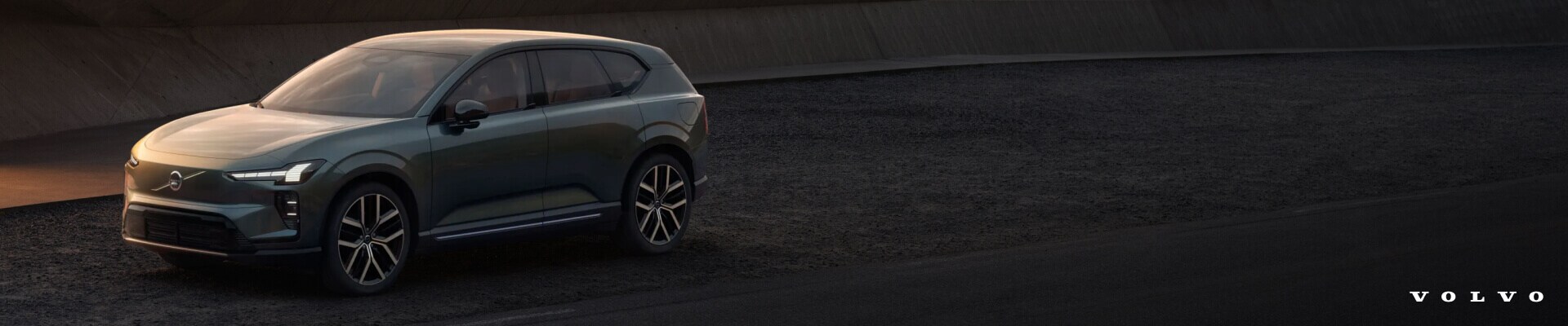 Front three-quarter view of a green Volvo EX60 Ultra on a dark ground next to a concrete structure.