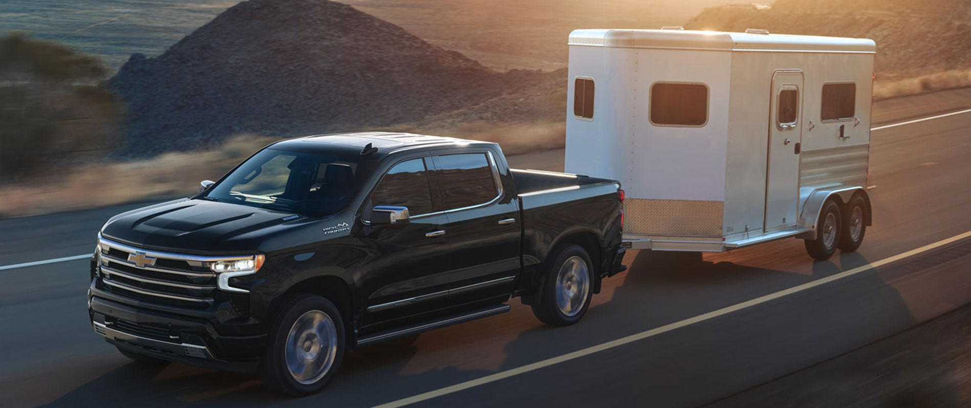 2026 Chevy Silverado 1500 Trim Comparison A black Chevy Silverado 1500 towing a trailer on the road above hills while the sun sets.