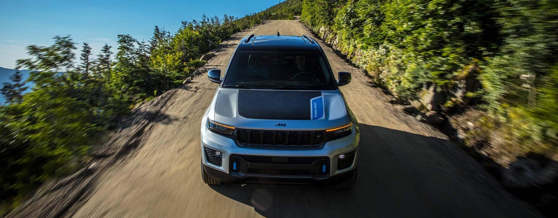 A front view of a silver Jeep Grand Cherokee driving down a dirt path on a hill. A front view of a silver Jeep Grand Cherokee driving down a dirt path on a hill.