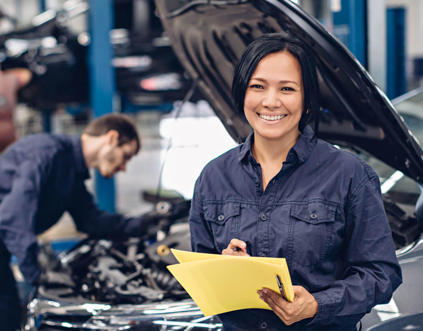 A mechanic holding a clipboard smiling while another mechanic works under the hood of a car in the background.
