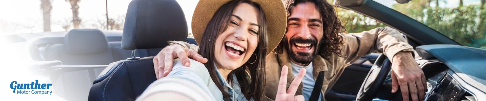 A couple smiles and makes a peace sign while taking a selfie in the front seat of a convertible.
