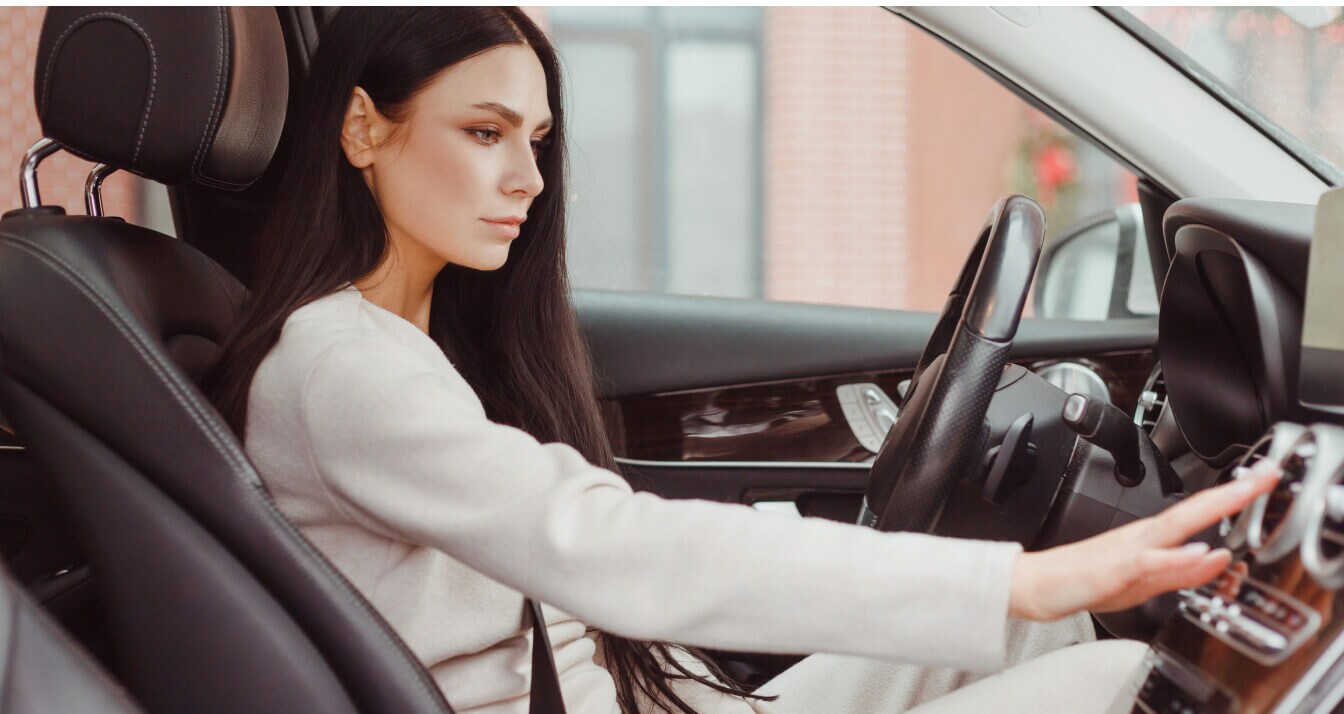 A concerned woman behind the wheel of a car holding her hand to her AC vents.