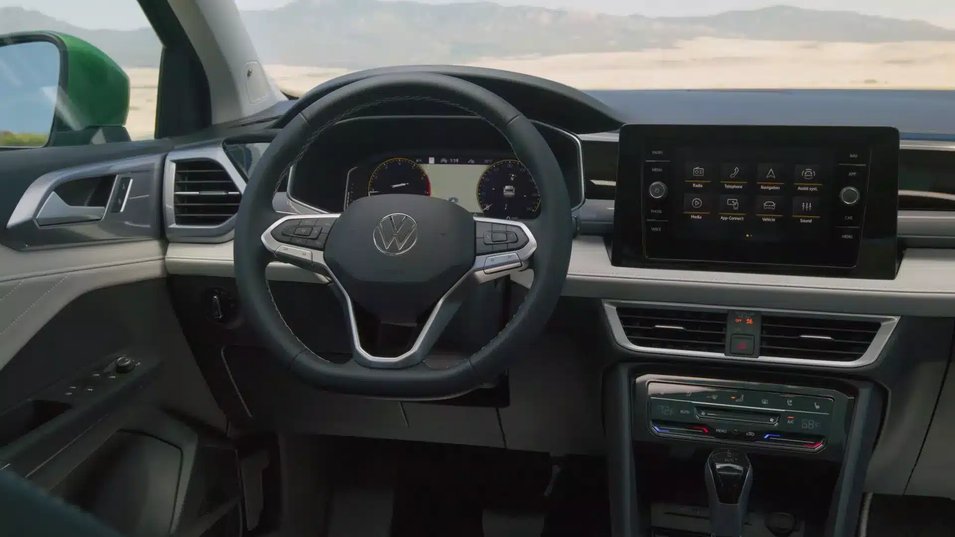 Interior of a new 2026 Volkswagen Taos, featuring a modern black and light gray dashboard. The steering wheel is multi-functional with a large central VW logo. The driver's view includes a digital instrument cluster and a large, horizontally-oriented touchscreen infotainment system located centrally above the climate controls. The overall setting suggests the car is parked outdoors in a dry, mountainous area.