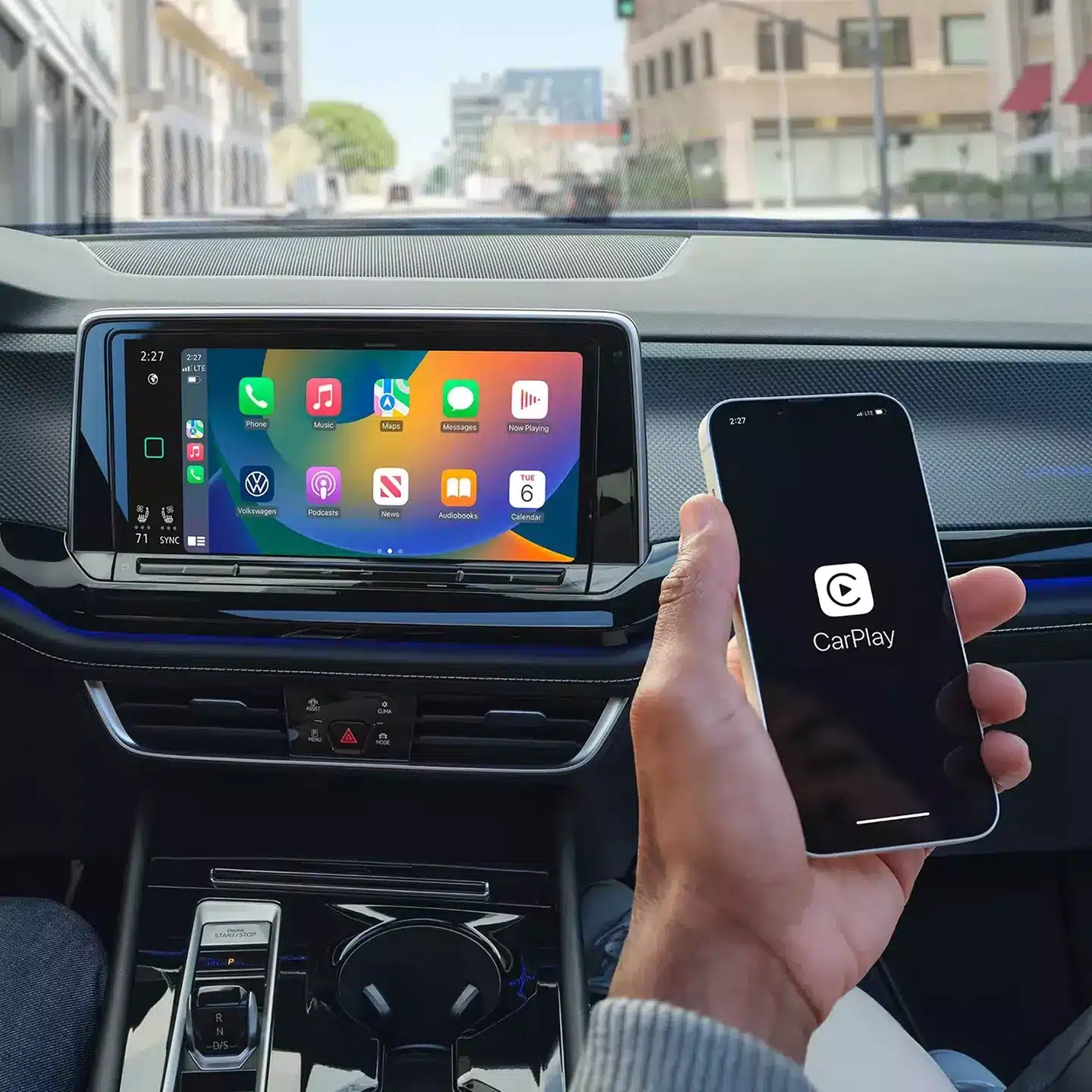 Hand holding a smartphone displaying the Apple CarPlay logo next to a car's dashboard screen showing the Apple CarPlay interface, set against an urban background.