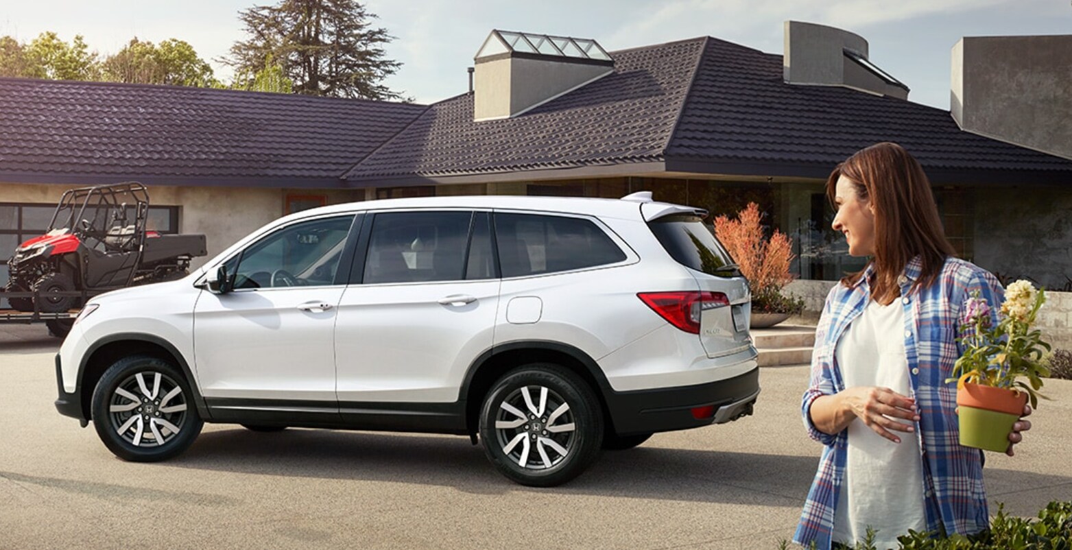 2022-Honda-Pilot-3.PNG A woman holding flowers looks back at the white 2022 Honda Pilot parked in her driveway