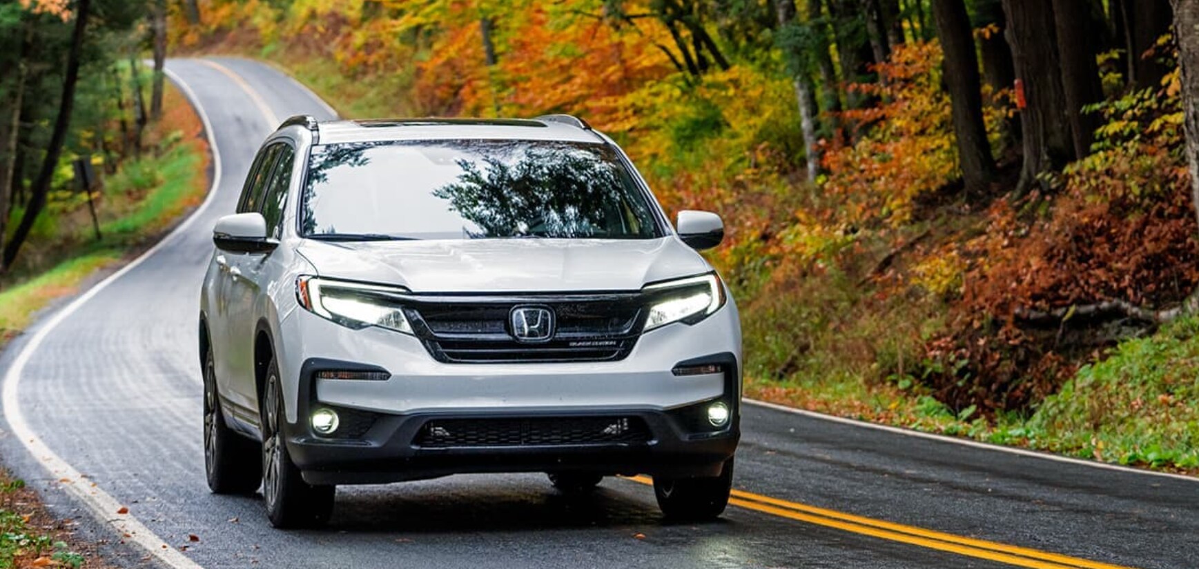 2022-Honda-Pilot-1.PNG A front view of a silver 2022 Honda Pilot as it cruises down a road in the woods