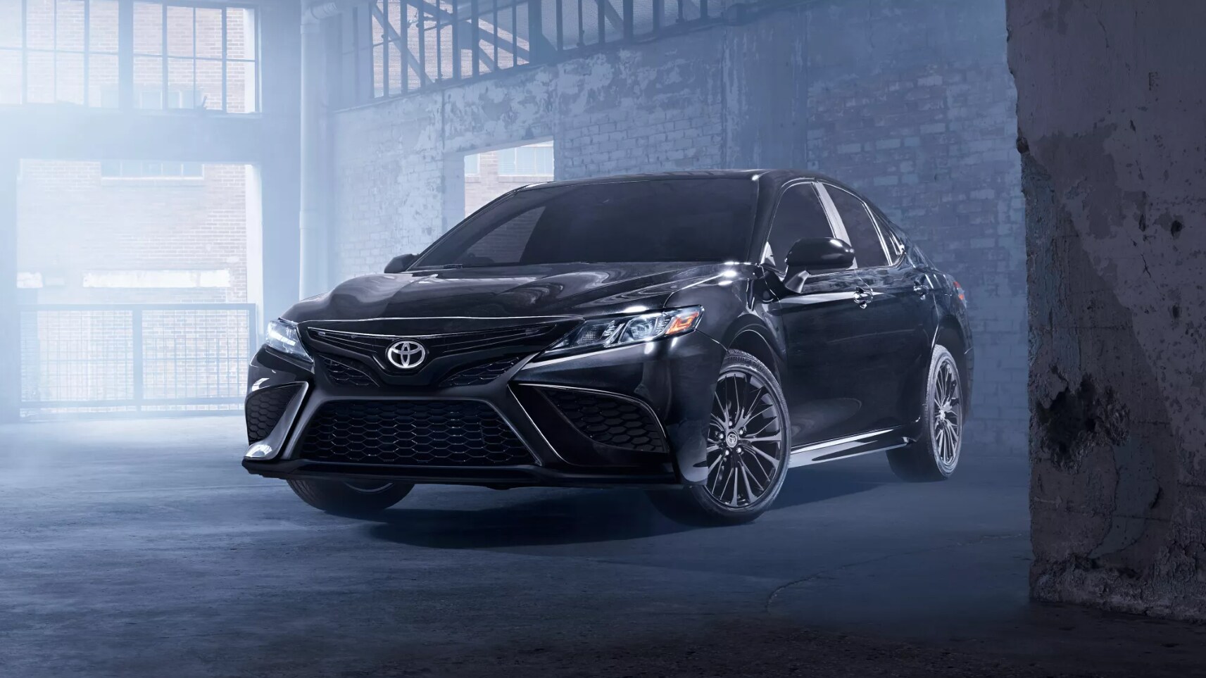 2022-Toyota-Camry-Hybrid-3.PNG A view of the front and profile of a black 2022 Toyota Camry Hybrid on a hazy morning parked in a warehouse