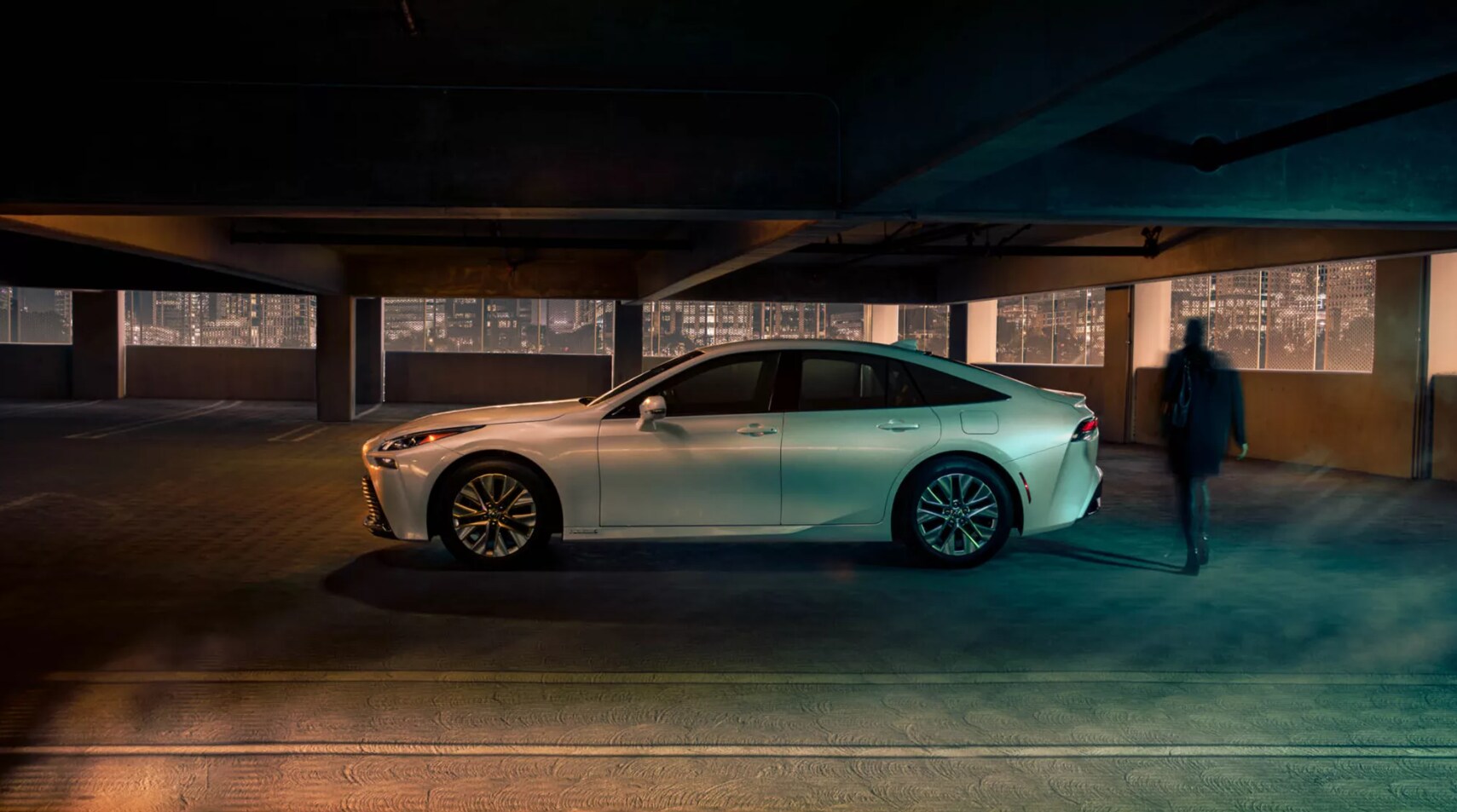 2022-Toyota-Mirai-3.PNG A profile view of a white 2022 Toyota Mirai as it sits parked at night in front of a cityscape