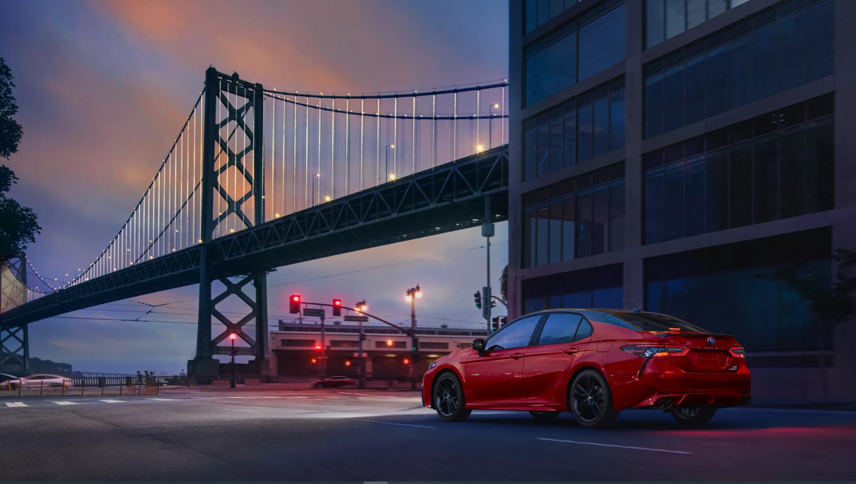 2022-Toyota-Camry-Hybrid-1.PNG A view of the rear and profile of a red 2022 Toyota Camry Hybrid as it drives down a street during dusk with the Golden Gate Bridge in the background