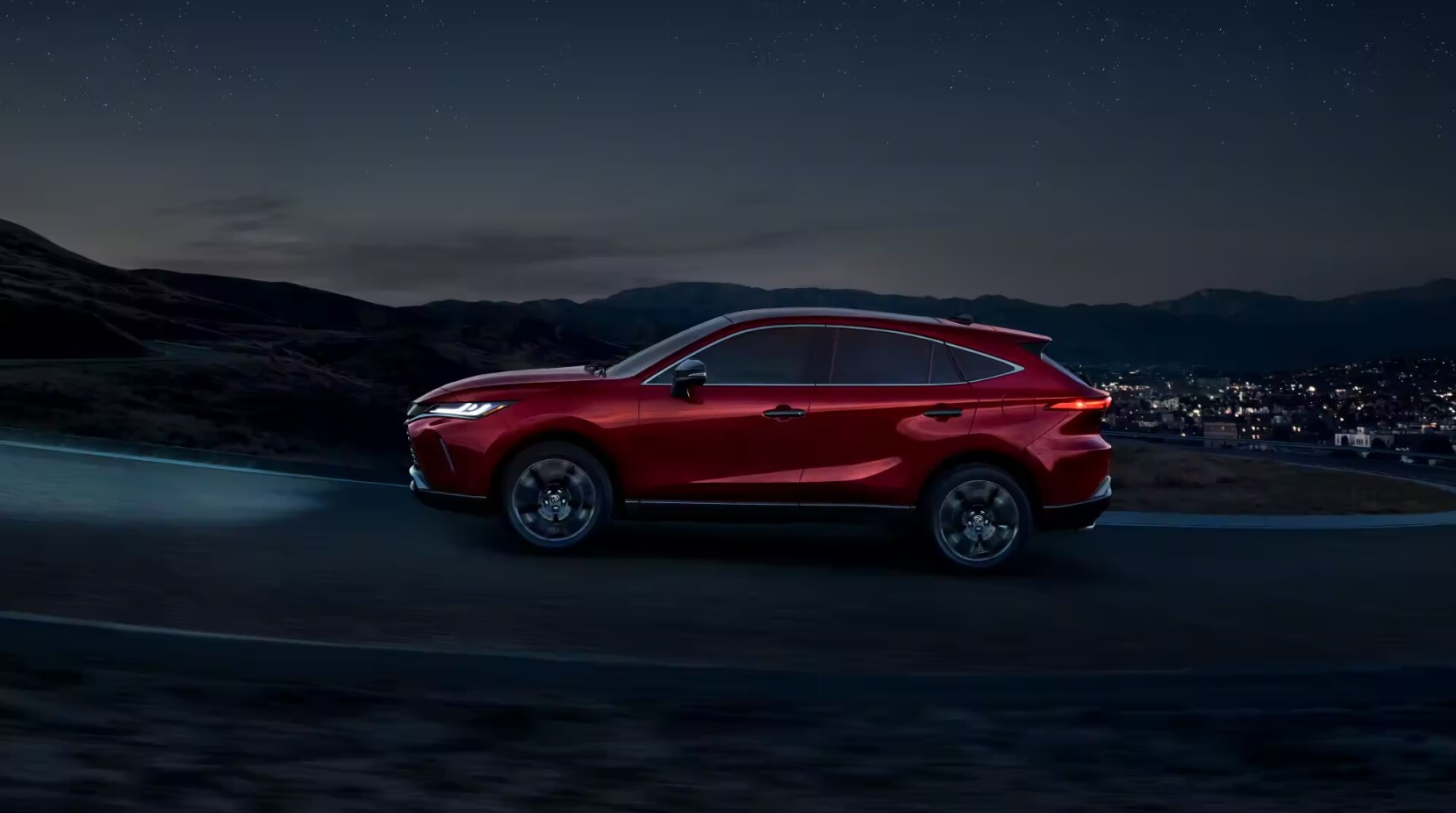 2023-toyota-venza-1.PNG A profile view of a red 2023 Toyota Venza Hybrid SUV driving on a highway at night