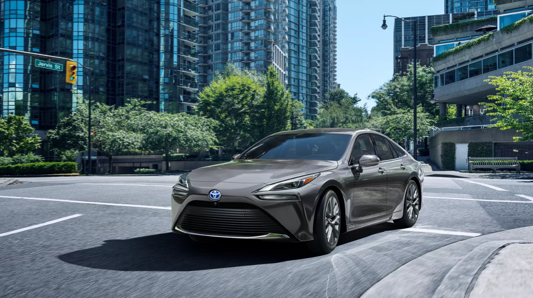 2022-Toyota-Mirai-1.PNG a smoke gray 2022 Toyota Mirai takes a left turn in a city intersection on a sunny day towards the viewer