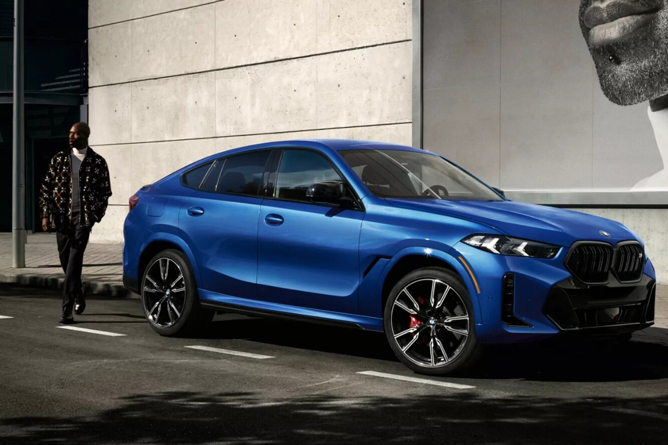 A man walking behind a blue BMW X6 parked by the curb in a city.