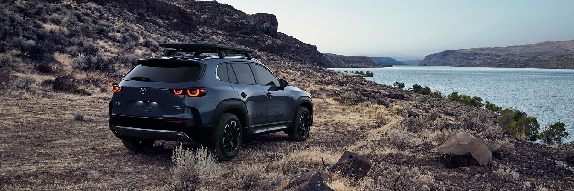 Model Features of the All-New 2023 Mazda CX-50 at Jack Giambalvo Mazda | Rear View of Black 2023 Mazda CX-50 Parked on Dirt and Rock Edge Looking Toward Water