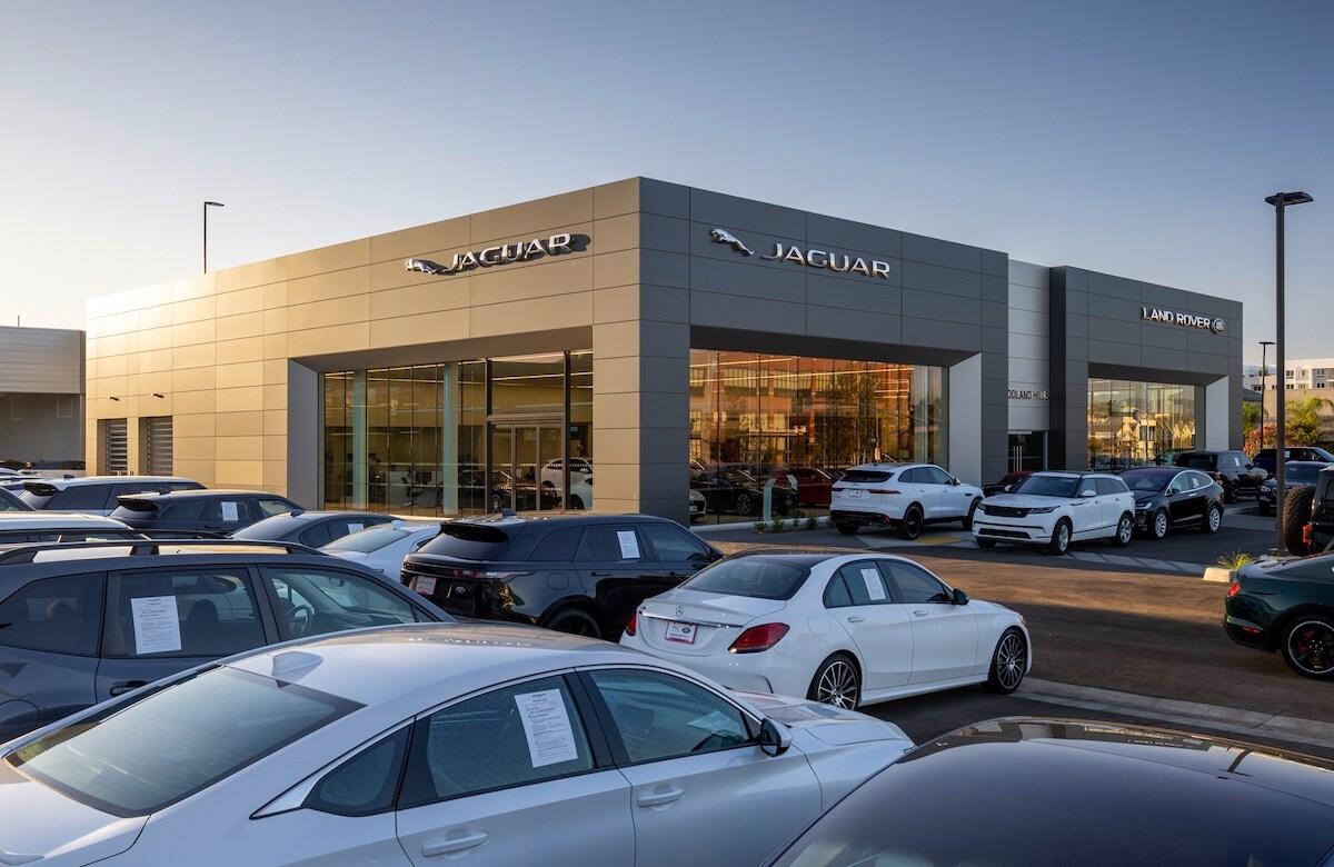 About Jaguar Woodland Hills, your Woodland Hills home for Jaguar vehicles. Image centered on Woodland Hills Jaguar Dealership building with the sun shining on the top left corner of the building. The sky is clear, and many vehicles are parked outside the building.