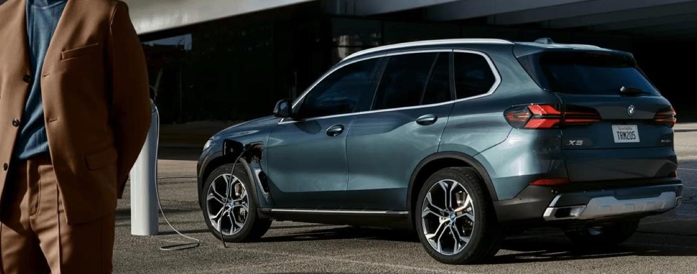 BMW store - 2023 BMW X5 - Blue Grey.jpg A man is shown walking near a blue 2023 BMW X5 parked on the side of a city street.