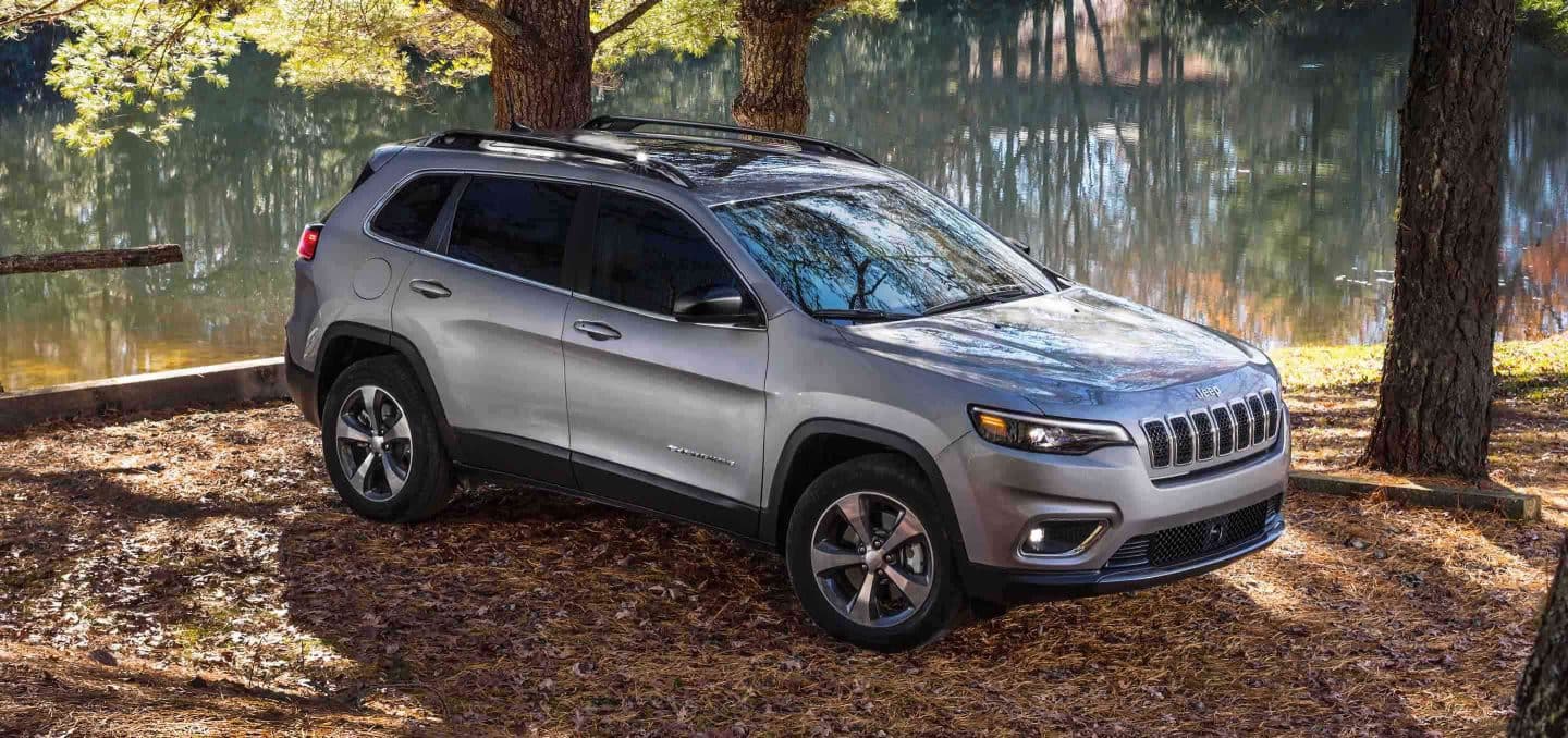 silver Jeep Cherokee SUV parked under some trees next to a stream