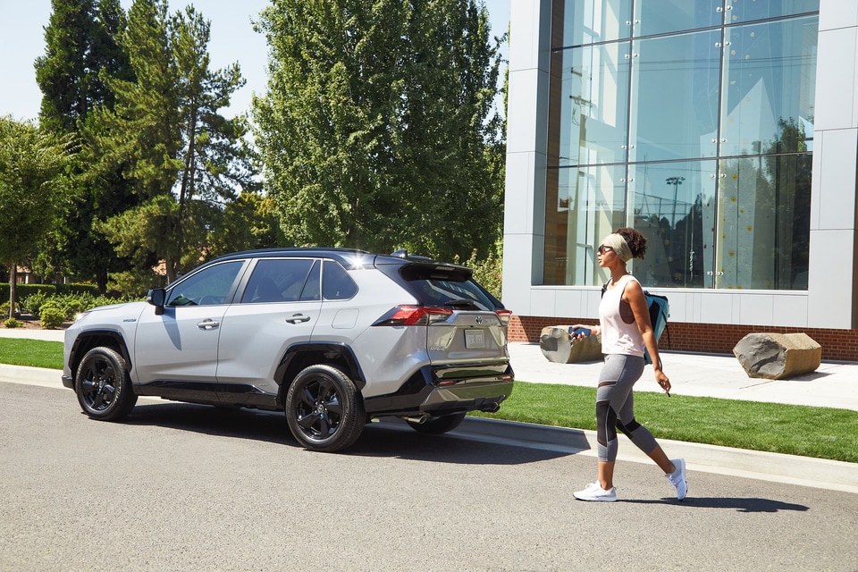 Silver RAV4 Hybrid parked in front of green trees and modern building as woman in athleisure clothes walks to the SUV