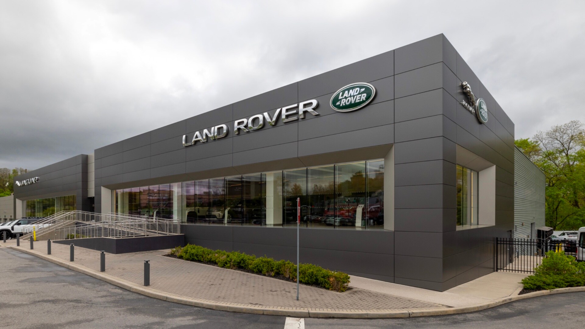 Grey Land Rover White Plains dealership building with a cloudy sky in the background. Vehicles can be seen inside through the large glass windows, and outside in the lot. A large LR logo and sign can be seen on the building.