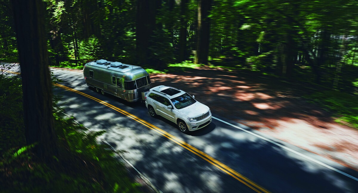 2022-Jeep-GrandCherokee-Summit-SUV-L04.png white Jeep Grand Cherokee SUV driving in a forested area, towing an airstream trailer